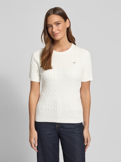 Modelka nosi Strickshirt mit Zopfmuster und Label-Stitching od Gant, mieszanka bawełny z dodatkiem streczu, krój slim fit, ścieg warkoczowy, wyhaftowane logo, okrągły dekolt, długość tyłu w rozmiarze S: 57 cm, długość rękawów w rozmiarze S: 24 cm, jasny kolor, widok całej sylwetki z przodu.