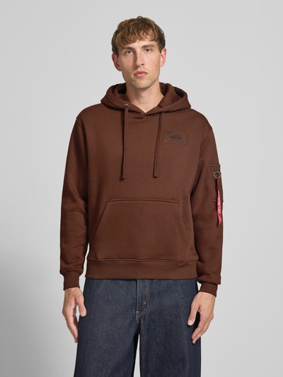 Männermodel trägt Hoodie mit Känguru-Tasche von Alpha Industries in Braun, Baumwoll-Mix, lockerer Schnitt, Ganzkörperansicht von vorne.