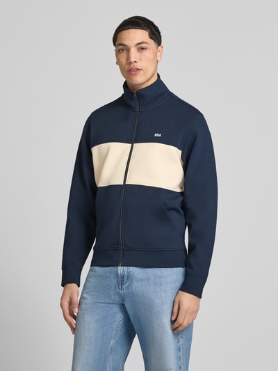 Męski model nosi bluzę rozpinaną z mieszanki bawełny z dodatkiem streczu od Helly Hansen, model 'KOSTER', prosty krój, stójka, zamek błyskawiczny, naszywka z logo, wpuszczane kieszenie, długość tyłu w rozmiarze M: 67 cm, długość rękawów: 66 cm, kolor granatowy z beżowym panelem, tekstura gładka, widok całej sylwetki z przodu.