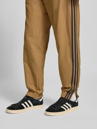 Mężczyzna model nosi Sweatpants mit elastischem Bund od adidas Originals w luźnym kroju, beżowy kolor, czarne paski po bokach, widok na nogi z przodu.