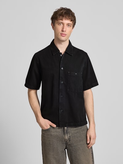 Mężczyzna model nosi Relaxed Fit Jeanshemd aus Lyocell-Baumwoll-Mix von Marc O'Polo Denim, czarny denim, krój relaxed fit, krótki rękaw, Kent kołnierz, zapięcie na guziki, widok całej sylwetki z przodu.