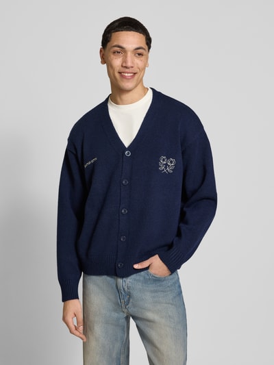 Männermodel trägt Cardigan mit Logo-Stitchings und Knopfleiste von ANOTHER COTTON LAB in Dunkelblau, Gerader-Schnitt, V-Ausschnitt, Gerippte-Abschlüsse, Rückenlänge 64 cm, Ärmellänge 59 cm, Ganzkörperansicht von vorne.