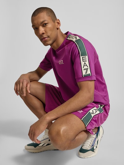 Męski model nosi T-shirt aus Viskose-Mix mit Logo od EA7 Emporio Armani, mieszanka wiskozy z dodatkiem streczu, krój regular fit, logo, okrągły dekolt, długość tyłu w rozmiarze M: 70 cm, mocny fiolet, akcenty bieli i zieleni, faktura gładka, widok całej sylwetki z przodu.