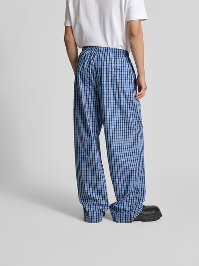 Mężczyzna model nosi piżamę spodnie z czystej bawełny od VETEMENTS w niebieską kratkę, krój relaxed fit, elastyczny pas, wpuszczane kieszenie, detal z logo, długość wewnętrzna nogawki w rozmiarze M: 86 cm, szerokość w rozmiarze M: 46 cm, zdjęcie całej sylwetki z tyłu.