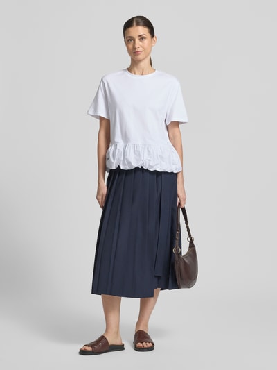 Frauenmodel trägt T-Shirt mit Rundhalsausschnitt von Rich & Royal aus reiner Baumwolle in Relaxed Fit, 1/2-Arm, Rückenlänge 59 cm, weiße Farbe, Einfachstruktur, kombiniert mit dunkelblauem Rock und braunen Accessoires, Ganzkörperansicht von vorne.