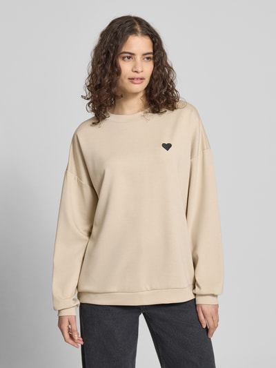 Vrouwenmodel draagt Relaxed Fit Pullover met hartborduursel model 'TALIA' van Pieces, effen beige, lange mouw, ronde hals, univormige structuur, vooraanzicht van het volledige lichaam.
