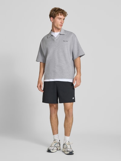 Męski model nosi szorty Relaxed Fit Shorts mit Logo-Stitching od New Balance, mieszanka bawełny, krój relaxed fit, wyhaftowane logo, elastyczny pas z tunelem, szerokość w rozmiarze M: 42 cm, długość wewnętrzna nogawki w rozmiarze M: 18 cm, widok całej sylwetki z przodu.