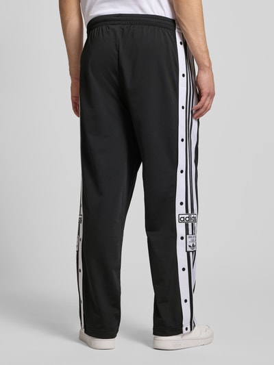 Männermodel trägt Trainingshose mit Label-Details und elastischem Bund von adidas Originals, Relaxed-Fit, Modell 'ADIBREAK', schwarze Hose mit weißen Streifen, Produktdetails laut Kontext, Ganzkörperansicht Rückansicht.