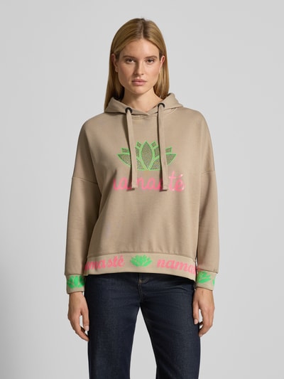 Frauenmodel trägt Oversized Hoodie mit Strasssteinbesatz Modell 'Namaste' von miss goodlife in Beige mit buntem Statement-Print, Ganzkörperansicht von vorne.