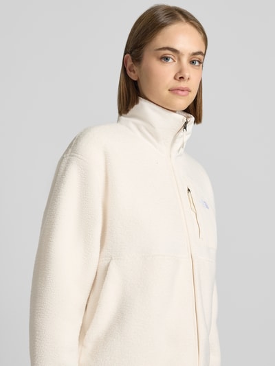 Kobieta nosi kurtkę z polaru o kroju regular fit Model 'YUMIORI' od The North Face w kolorze ecru, stójka, wyhaftowane logo, zamek błyskawiczny, Polar. Szerokość w rozmiarze S: 52 cm, Szerokość klatki piersiowej w rozmiarze S: 58 cm, Długość tyłu w rozmiarze S: 58 cm, Długość rękawów w rozmiarze S: 57 cm. Zbliżenie na górną część ciała.