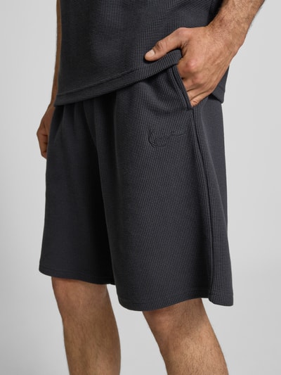 Männermodel trägt Relaxed Fit Shorts mit Strukturmuster von KARL KANI aus Baumwoll-Mix in Dunkelgrau, Nahaufnahme Oberschenkel.
