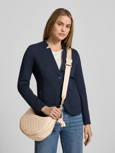 Frauenmodel trägt Handtasche mit Label-Applikation Modell 'cortina 1.0 jennifer' von JOOP! mit Logo-Muster, Schulterriemen, Hauptfach mit Reißverschluss, beige Farben und sichtbarer Textur, Ganzkörperansicht von vorne.