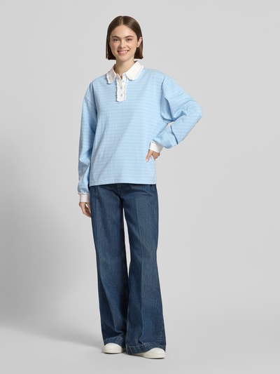 Frauenmodel trägt Relaxed Fit Poloshirt mit Label-Stitching Modell 'Jolina' von OH APRIL aus reiner Baumwolle mit Spitzenbesatz und Knopfleiste in Hellblau, kombiniert mit Jeans, Ganzkörperansicht von vorne.