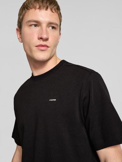Mężczyznę model nosi Regular Fit T-Shirt mit Logo-Print Modell 'True' von G-Star Raw, czarny, okrągły dekolt, długość tyłu w rozmiarze M: 66 cm, zbliżenie na górną część ciała