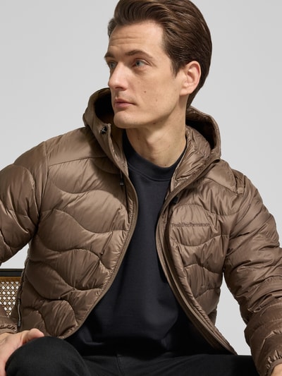 Męski model nosi Regular Fit Jacke mit Logo-Stitching Modell 'Helium' od Peak Performance w odcieniu brązowym; krój regular fit, pikowana tekstura, kaptur ze ściągaczem, zamek błyskawiczny, kieszenie na zamek, elastyczne wykończenia; długość tyłu w rozmiarze M: 72 cm, szerokość ramion: 49 cm, szerokość klatki piersiowej: 56 cm, długość rękawa: 69 cm; widok z przodu półzbliżenie.