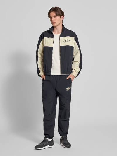 Männermodel trägt Trainingshose mit Logo und elastischem Bund von Nike, Relaxed-Fit, dunkle Farben, Ganzkörperansicht von vorne. Innenbeinlänge bei Größe M: 76 cm, elastische Beinabschlüsse, Logo.
