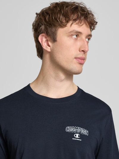 Männermodel trägt T-Shirt mit Logo und Rundhalsausschnitt von CHAMPION in Dunkelblau, reine Baumwolle, Regular Fit, Nahaufnahme Oberkörper.