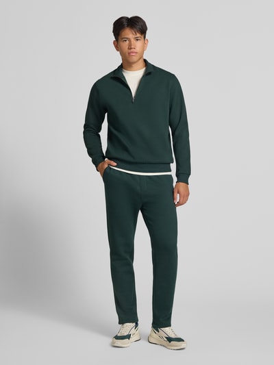 Männermodel trägt Regular Fit Sweatshirt mit Baumwoll-Anteil Modell 'Bradley' von JACK & JONES in Dunkelgrün, Ganzkörperansicht von vorne, Material-Mix mit Baumwoll-Anteil, Troyer-Kragen, Rückenlänge 68 cm, Ärmellänge 66 cm.