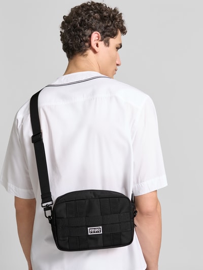 Männermodel trägt Crossbody Bag mit Logo-Stitching von Kenzo in Schwarz mit Strukturmuster, Steckfach innen, Abnehmbarer Schulterriemen, Ganzkörperansicht Rückansicht.