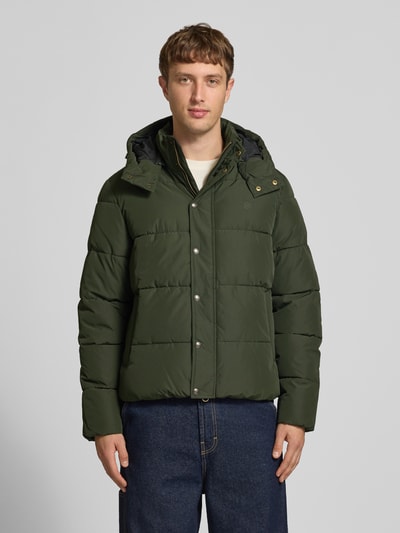 Männermodel trägt Steppjacke mit Kapuze und Reißverschluss von Superdry in Dunkelgrün, sichtbare Steppnähte, Ganzkörperansicht von vorne. Regular Fit, Stehkragen, verdeckter Reißverschluss, Knopfleiste, Rückenlänge 71 cm, Schulterbreite 50 cm, Brustweite 56 cm, Ärmellänge 83 cm.