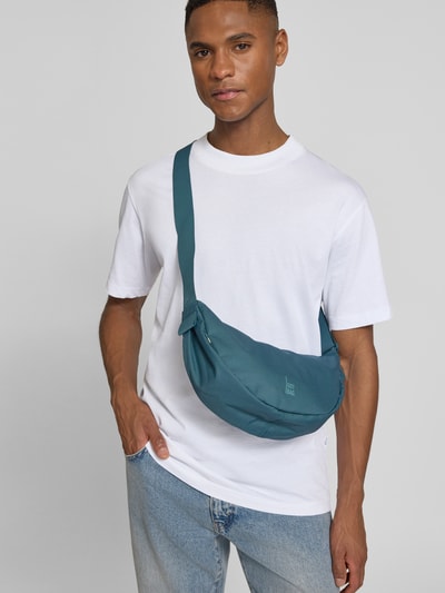 Männermodel trägt Umhängetasche mit Label-Detail von Gotbag in Petrol, Reißverschluss, verstellbarer Schulterriemen, Ganzkörperansicht von vorne. Kontext: Herren Umhängetasche, Hauptfach mit Reißverschluss, Innenfach mit Reißverschluss, Breite 31 cm, Länge 19 cm, Tiefe 9 cm, Länge Schulterriemen 87 cm.