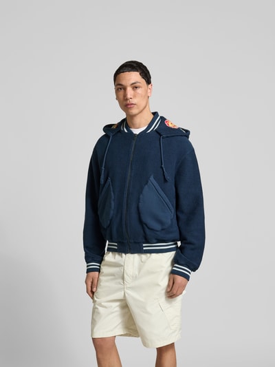 Männermodel trägt Relaxed Fit Jacke mit Motiv-Patches von Kenzo, dunkelblau, Baumwolle, aufgesetzte Taschen, Reißverschluss, Ganzkörperansicht von vorne.