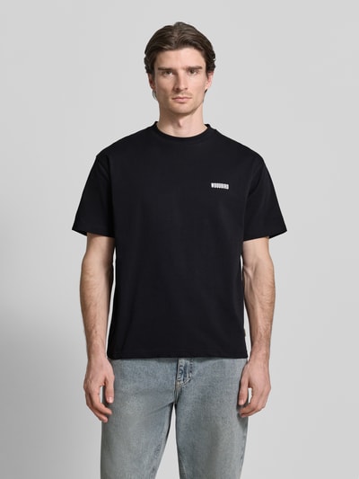 Męski model nosi T-shirt mit Label-Print Modell 'Baine' von Woodbird, czarny, bawełna, krój regular fit, okrągły dekolt, nadruk z logo, długość tyłu w rozmiarze M: 67 cm, Ganzkörperansicht von vorne.