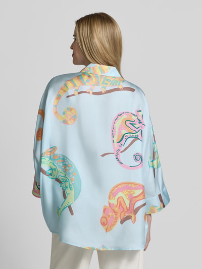 Frauenmodel trägt Bluse mit Hemdblusenkragen und 3/4-Arm von HERZENS, hellblau mit bunten Chamäleon-Motiven, Ganzkörperansicht Rückansicht.