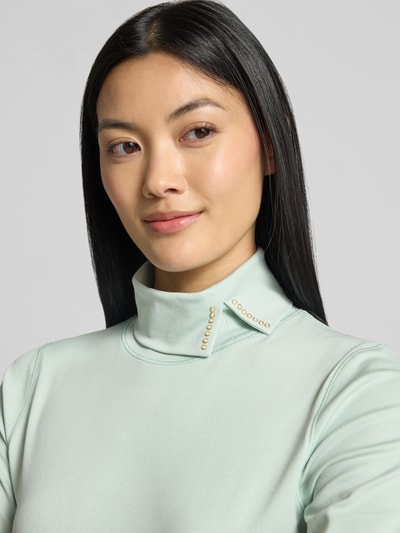Vrouwenmodel draagt Slim Fit T-Shirt uit katoenmix van Marc Clain, asymmetrische col, siersteentjes, kleur lichtgroen, Nahaufnahme bovenlichaam, Ruglengte bij maat 36: 56 cm.