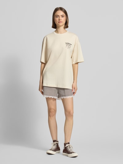 Frauenmodel trägt Oversized T-Shirt mit Statement-Print von REVIEW aus reiner Baumwolle im Oversize Fit, Rundhalsausschnitt, Farbe Beige, kombiniert mit gemusterten Shorts, Rückenlänge bei Größe S: 70 cm, Ganzkörperansicht von vorne