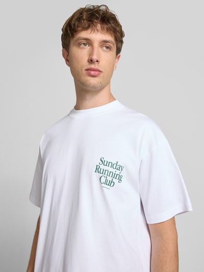 Männermodel trägt Herren T-Shirt mit Statement-Print Modell 'Sunday Running Club' von ANOTHER COTTON LAB, reine Baumwolle, Boxy-Fit, Rundhalsausschnitt, weiß mit grünem Aufdruck, Ganzkörperansicht von vorne.