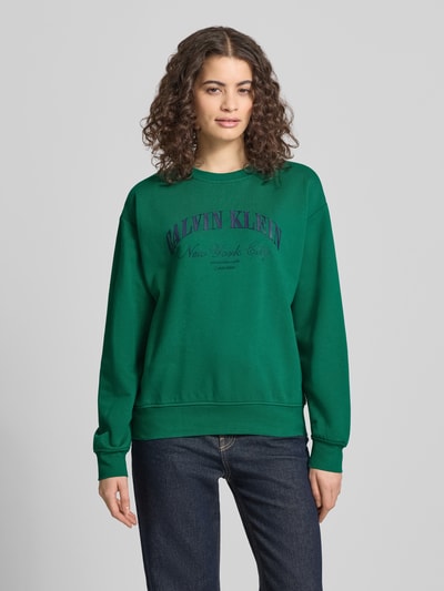 Frauenmodel trägt Sweatshirt mit Label-Print Modell 'GRAPHIC' von Calvin Klein Jeans in Grün mit Rundhalsausschnitt und elastischen Abschlüssen, Baumwoll-Elasthan-Mix, lockere Passform, Ganzkörperansicht von vorne.