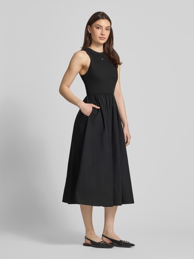 Frauenmodel trägt Midikleid aus reiner Baumwolle von Tommy Jeans, unifarben schwarz, ärmellos, A-Linie, Ganzkörperansicht von vorne.