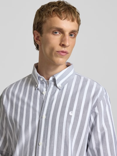 Männermodel trägt Regular Fit Freizeithemd mit Logo-Stitching Modell 'Deven' von Carhartt Work In Progress, Reine Baumwolle, Langärmelig, Button-Down-Kragen, durchgängige Knopfleiste, Sportmanschetten, vertikale weiße und graue Streifen, weiche Textur, Nahaufnahme Oberkörper.