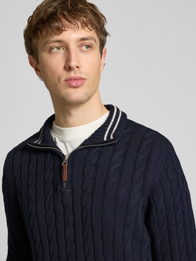 Männermodel trägt Strickpullover aus reiner Baumwolle von Superdry in Dunkelblau mit Zopfmuster, Troyer-Kragen, Nahaufnahme Oberkörper.