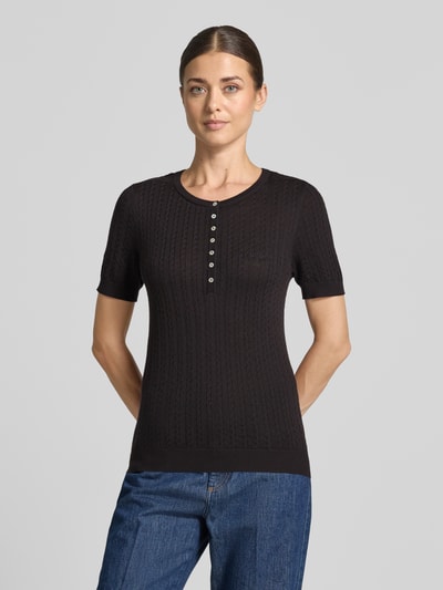 Frauenmodel trägt Wollpullover aus reiner Wolle Modell 'MEADIE' von Cph Muse, Schwarz, Zopfmuster, Knopfleiste, Kurzarm, Ganzkörperansicht von vorne.