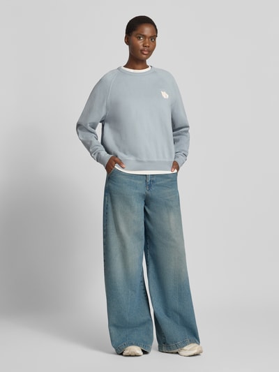 Vrouwenmodel draagt Relaxed Fit Sweatshirt aus reiner Baumwolle mit Statement-Print van ba&sh, lichtblauw, puur katoen, relaxed fit, ribboorden, lange mouw, statementprint, raglanmouwen, dames sweatshirt, Ganzkörperansicht van voren.