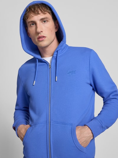 Männermodel trägt Sweatjacke mit Kapuze und Logo-Stitching von Superdry aus Baumwoll-Mix in Blau, seitliche Eingrifftaschen, Nahaufnahme Oberkörper.