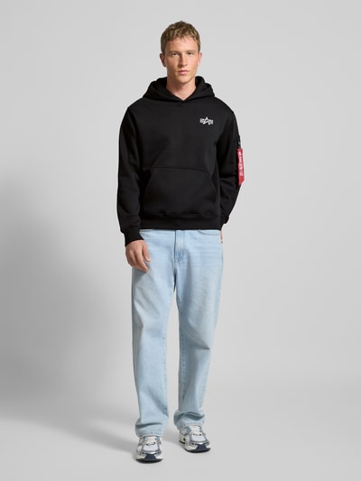Männermodel trägt Hoodie mit Label-Details von Alpha Industries in Schwarz, Baumwoll-Mix, Känguru-Tasche, Lockerer-Schnitt, Ganzkörperansicht von vorne.
