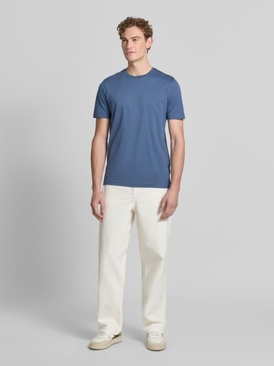 Męski model nosi T-Shirt aus Baumwolle mit Rundhalsausschnitt von Christian Berg Men, krój slim fit, okrągły dekolt, długość tyłu 73 cm w rozmiarze m, stalowoniebieski, prosta faktura, gładka powierzchnia, jasne spodnie, całopostaciowy widok z przodu.