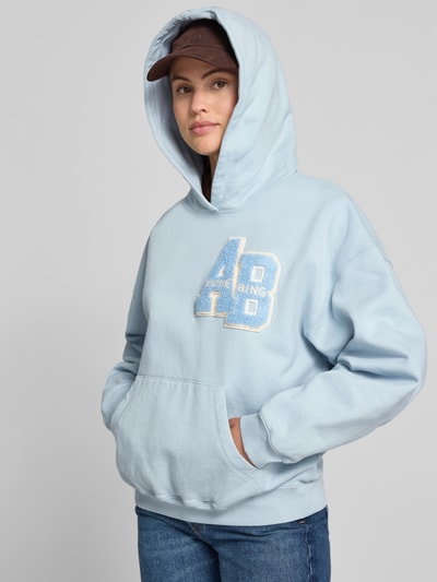 Frauenmodel trägt Relaxed Fit Hoodie mit Logo-Detail von Anine Bing aus reiner Baumwolle in Hellblau mit Känguru-Tasche, elastische Abschlüsse, Rückenlänge bei Größe S 85 cm, Ganzkörperansicht von vorne.