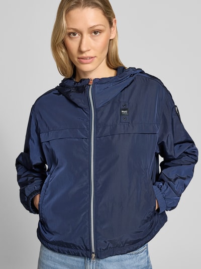 Frauenmodel trägt Trekking Jacke mit Kapuze Modell 'BURRAGE' von Blauer Usa in Dunkelblau, glatte Struktur, Nahaufnahme Oberkörper. Gerader Schnitt, Zweiwege-Reißverschluss, Label-Patch und seitliche Reißverschlusstaschen ergänzen das Design. Elastische Ärmelabschlüsse, Rückenlänge 56 cm, Schulterbreite 48 cm, Brustweite 56 cm, Ärmellänge 57 cm.