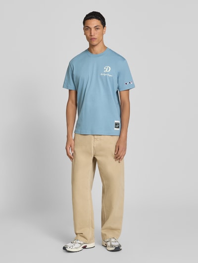 Männermodel trägt Relaxed Fit T-Shirt aus reiner Baumwolle von Champion in Hellblau mit Statement-Print, ganzkörperansicht von vorne.