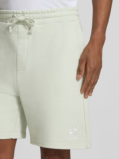 Mężczyzna nosi Regular Fit Sweatshorts aus reiner Baumwolle Modell 'DARONOSO' von HUGO, efekt: jednolity kolor, krój regular fit, wyhaftowane logo, ozdobne szwy, ściągacz z tunelem, wpuszczane kieszenie, kieszeń z tyłu, wykończenie materiału: dzianina dresowa, bawełna, zbliżenie na górną część nogi od przodu.