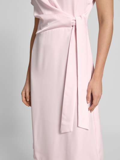 Frauenmodel trägt Knielanges Cocktailkleid mit Knotendetail und Reißverschluss von Rinascimento in zartem Rosa, Nahaufnahme Oberkörper; Taillierter Schnitt, Rundhalsausschnitt, Knielänge, Rückseitiger Reißverschluss, Rückenlänge 105 cm, Brustweite 46 cm, Ärmellänge 17 cm.