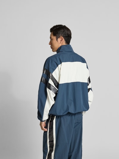 Männermodel trägt Trainingsjacke mit Stehkragen von VETEMENTS in Blau und Weiß, Relaxed-Fit, elastische Abschlüsse, Rückenlänge 74 cm, Zweiwege-Reißverschluss, Schulterbreite 50 cm, Ärmellänge 73 cm, Eingrifftaschen, Ganzkörperansicht Rückansicht.