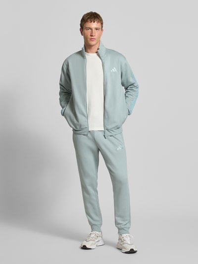 Männermodel trägt Sweatanzug mit Label-Stitchings von adidas Sportswear in Hellblau, Baumwoll-Mix, Regular-Fit, Ganzkörperansicht von vorne.