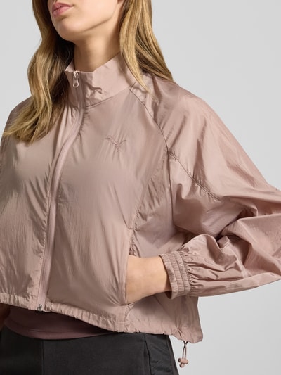Frauenmodel trägt Relaxed Fit Jacke mit Stehkragen Modell 'Her' von PUMA in Hellrosa, Logo-Stitching, Reißverschluss, Eingrifftaschen, elastische Ärmelabschlüsse, leichte strukturierte Textur, Nahaufnahme Oberkörper. Rückenlänge 55 cm, Schulterbreite 45 cm, Brustweite 56 cm, Ärmellänge 64 cm bei Größe S.