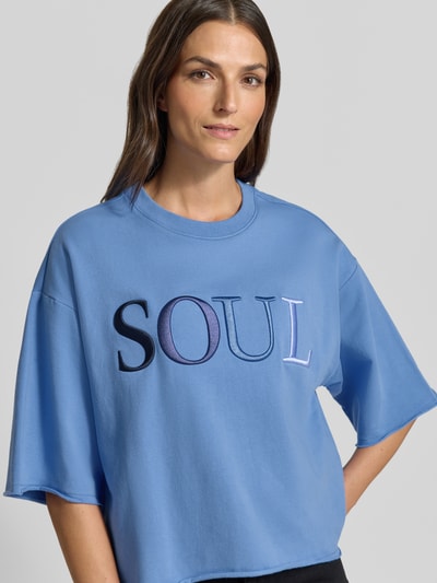 Frauenmodel trägt Cropped Boxy Fit Sweatshirt mit Label-Stitching von Smith and Soul in Hellblau, Rundhalsausschnitt, 1/2-Arm, Nahaufnahme Oberkörper. Baumwoll-Elasthan-Mix mit weicher Textur, dezentes Farbspiel im Logo.