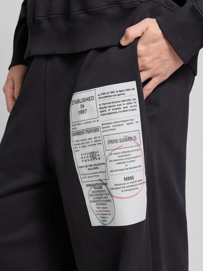 Männermodel trägt Loose Fit Stoffhose mit Logo-Print von MM6 Maison Margiela in Schwarz, Baumwolle, seitliche Taschen, Nahaufnahme Oberschenkel.